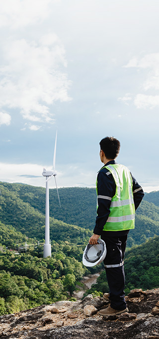 Turbine technician in neon safety jacket standing on the top of the hill and looking at the perspective of hills covered with greenery and wind turbines above them.